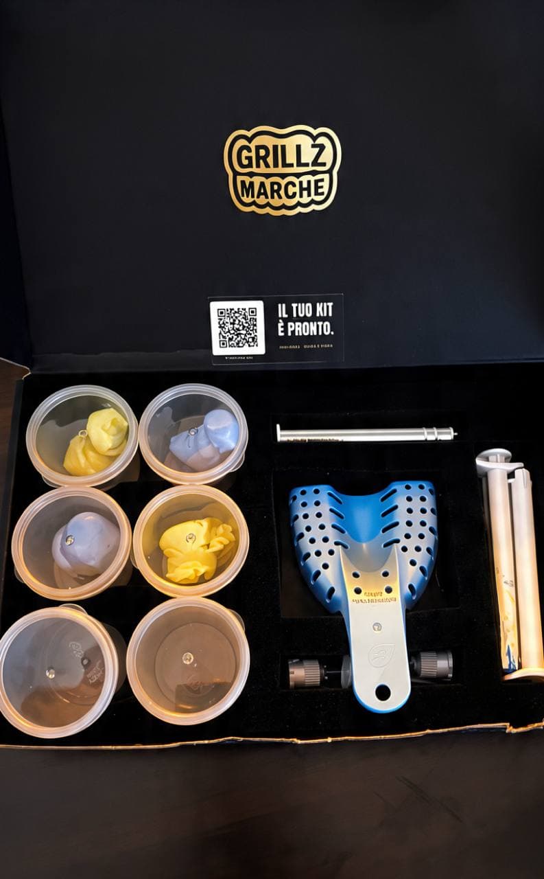 Kit impronta Grillz Marche aperto: scatola nera col logo oro, sticker «Il tuo kit è pronto» col QR della video-guida, sei barattolini di pasta pre-dosata, porta-impronte blu, siringa a doppia camera coi puntali e spatola per miscelare