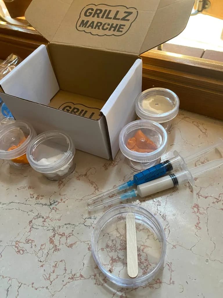 Contenuto reale del kit impronta: scatola timbrata Grillz Marche aperta, barattolini di pasta pre-dosata, due siringhe di pasta fluida con puntali e vassoietto col bastoncino di legno